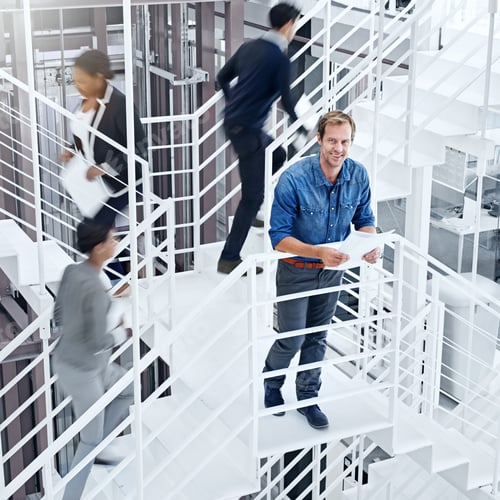 Preview: Portrait of a young professional standing on a stairs with colleagues rushing around him