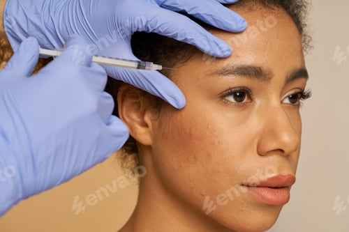 Preview: Portrait of beautiful young woman looking away while getting face injections isolated over beige