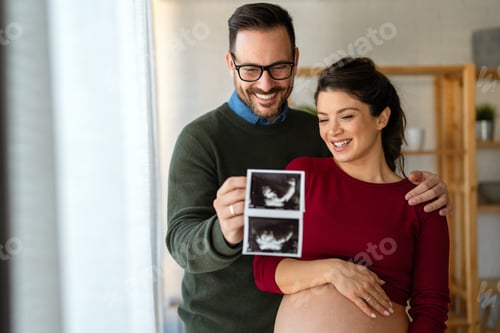 Preview: Happy young couple, pregnant woman, holding and showing ultrasound scan picture at home