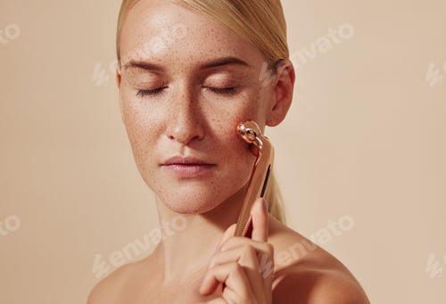 Preview: Female with smooth freckled skin using face roller against a beige background.