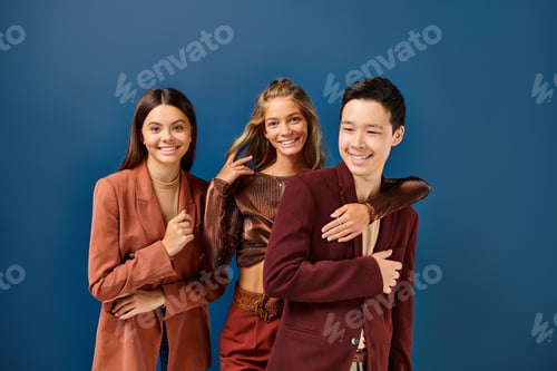 Preview: happy diverse teenagers in stylish vibrant attires posing together on blue backdrop, friendship day