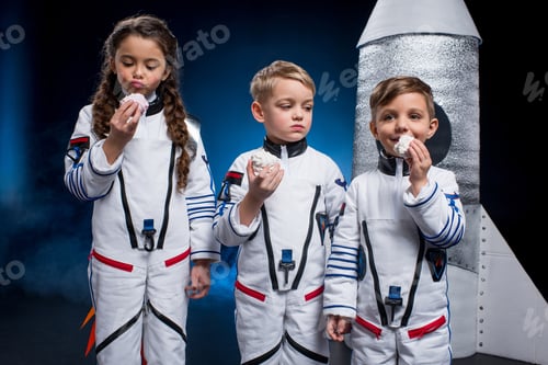 Preview: Three cute kids in astronaut costumes eating sweets standing near rocket