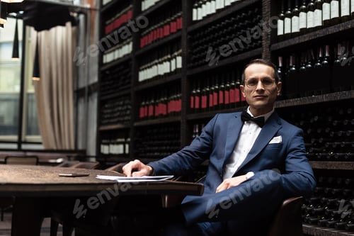 Preview: happy handsome man in stylish suit sitting in armchair in front of wine storage