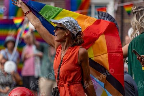 Preview: Old woman in red dress rising the rainbow flag in the crowd during the canal pride 2024