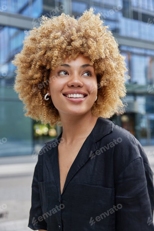 Preview: Vertical shot of positive curly haired woman dressed in casual black shirt smiles happily looks some