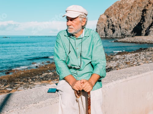 Preview: Portrait of a melancholic retired senior man sitting near the beach with hands on walking stick