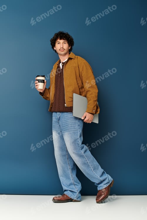 Preview: Stylish young man stands confidently holding coffee and a laptop against a bold blue backdrop