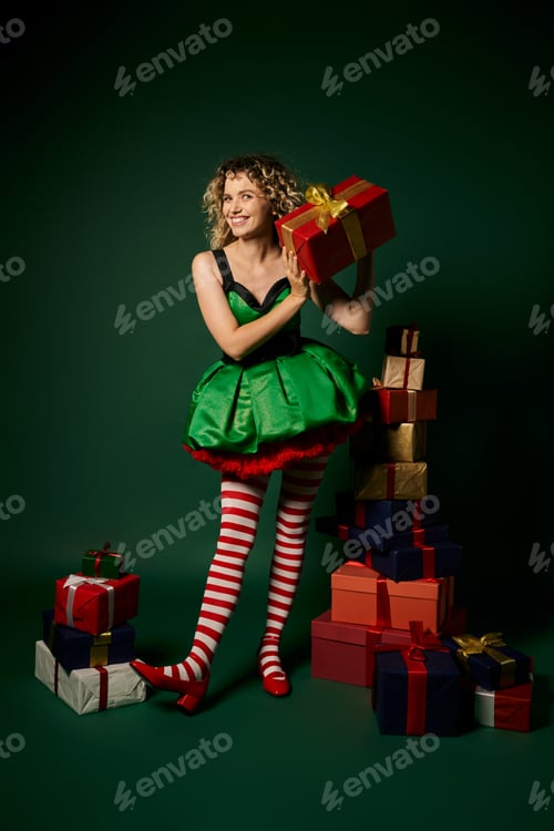 Preview: curly new year elf standing near pile of gifts and mysteriously showing one of them at camera