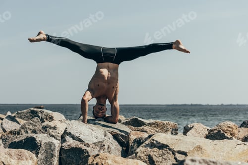 Preview: handsome shirtless man practicing yoga on rocks and performing tripod headstand in front of sea