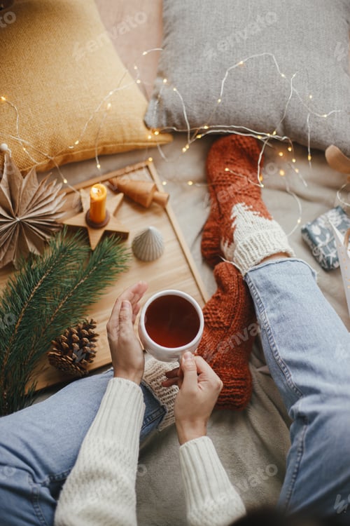 Preview: Woman hands with warm cup of tea on soft bed with christmas stars, lights, candle. Cozy winter home