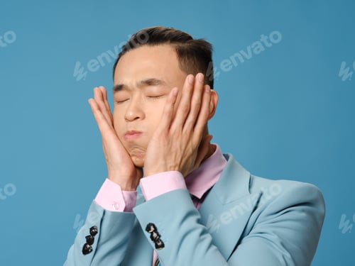 Preview: Asian man with puffy cheeks touching face with hands on blue background cropped view