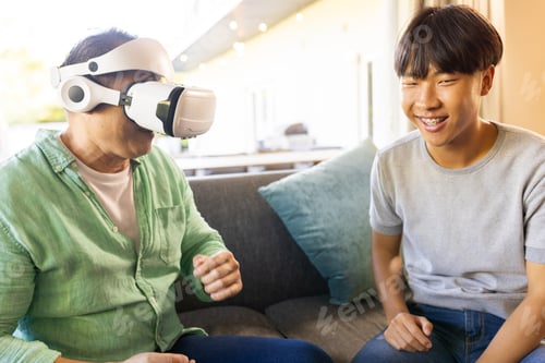 Preview: Using VR headset, asian father sitting on couch with smiling teenager at home