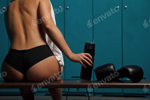 Preview: Close-up pretty young woman sit with an athletic with water bottle and Boxing gloves in locker room