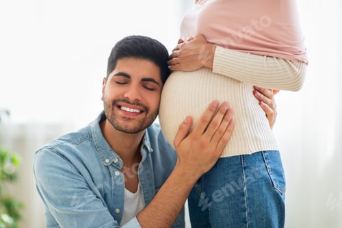 Preview: Delighted eastern man embracing tummy of his pregnant wife, listening baby kicks, tenderly touching