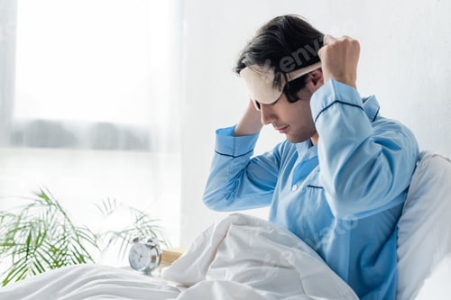 Preview: man in blue pajamas sitting on bed in morning and taking off sleep mask