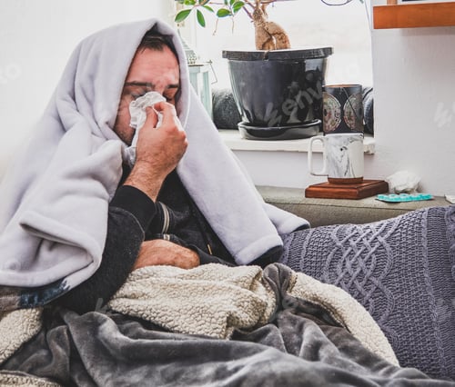 Preview: Sick young man sitting sitting on couch and blowing nose.