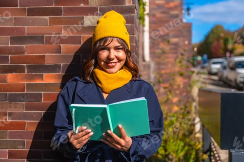 Preview: Young woman smiling reading book during urban autumn day