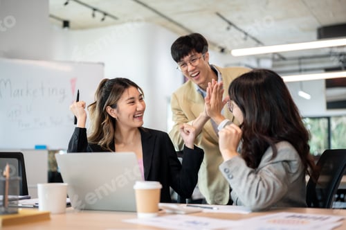 Preview: Asian businesswoman and coworker looking excited while raising a fist and clapping hands in office.