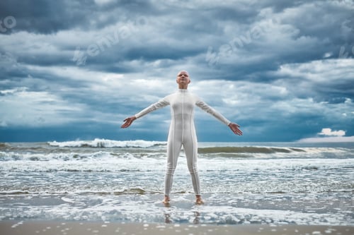 Preview: Happy hairless girl with alopecia in white futuristic suit stands on beach bathed by ocean waves