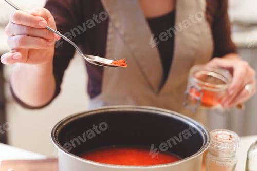 Preview: Female cook adds paprika spices while cooking tomato soup.Close-up view on spoon with red pepper.