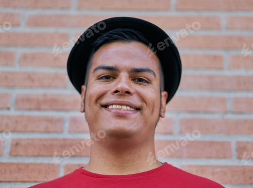 Preview: Closeup shot of the smiling face of a young male in a fedora hat in front of a cobblestone wall
