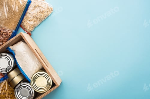 Preview: top view of cans and groats in zipper bags in wooden box on blue background, food donation concept