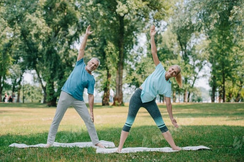 Preview: smiling mature man and woman practicing warrior yoga poses while standing on yoga mats in park