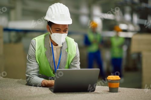 Preview: Black warehouse manager working on laptop and wearing face mask due to coronavirus.