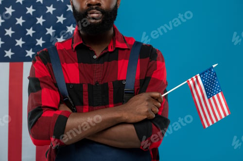 Preview: Young bearded repairman of African ethnicity in workwear holding American flag