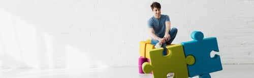 Preview: panoramic shot of cheerful man looking at blue and green jigsaw pieces while sitting near brick wall
