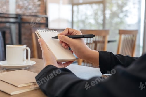 Preview: Closeup image of a hand holding and writing on blank notebook with coffee cup on table in cafe