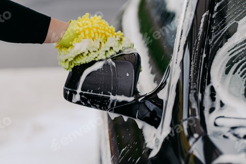 Preview: Mans hand holding sponge for washing car. Black auto with soap bubbles