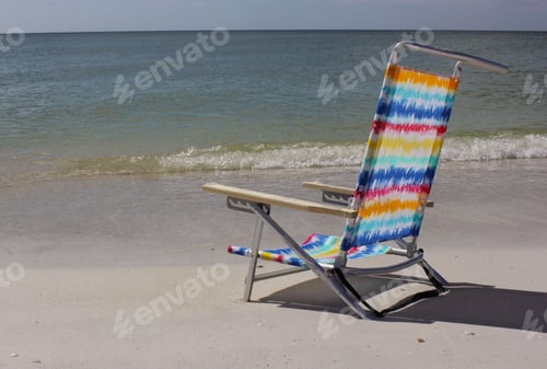 Preview: Colorful beach chair on sandy shore with waves.