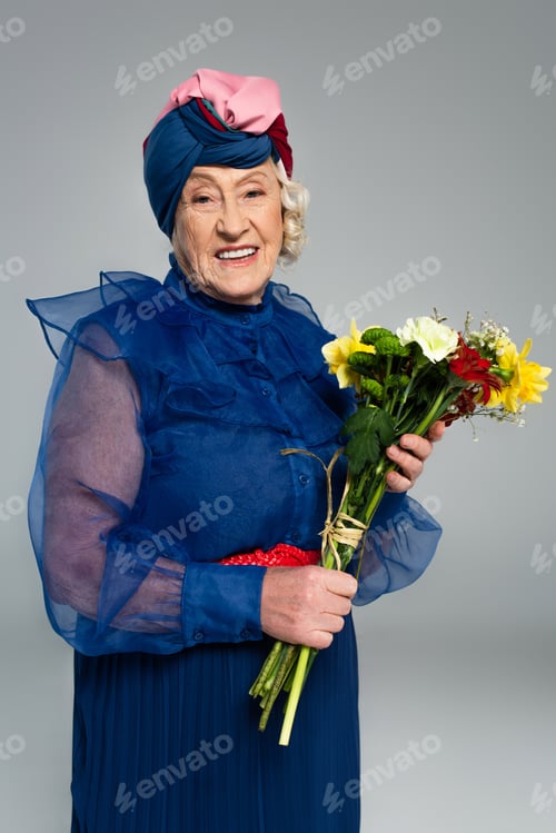 Preview: smiling elderly woman in blue dress and turban holding bouquet of flowers isolated on grey