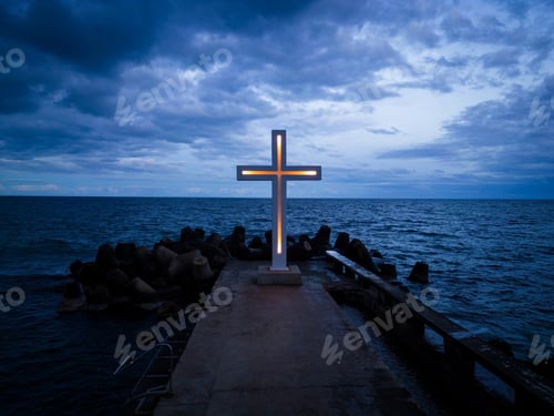 Preview: A large Christian cross stands at the edge of a pier against a dramatic sky and sea, seen from above