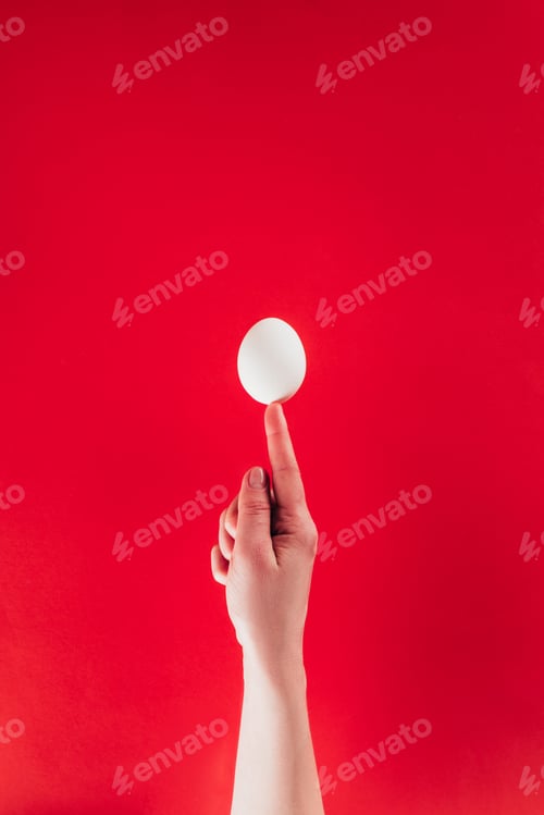 Preview: partial view of woman holding raw chicken egg on finger isolated on red