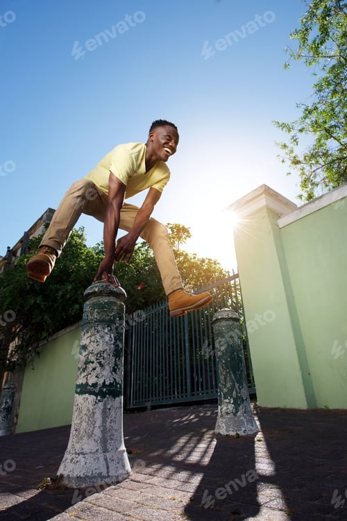 Preview: happy young african man jumping over iron post on sidewalk