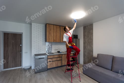 Preview: Young man installing ceiling lamp on stepladder in kitchen