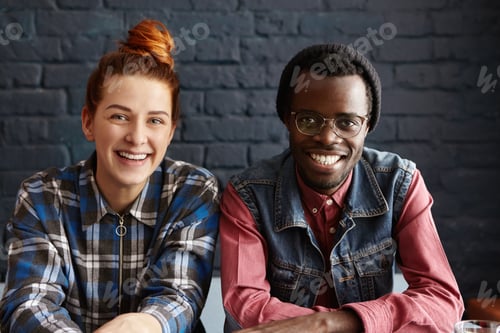 Preview: Cute interracial couple relaxing indoors, sitting against black brick wall background. Two stylish y