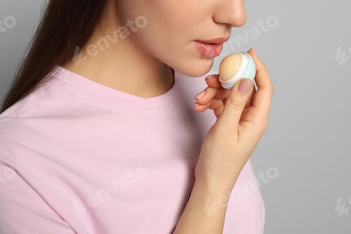 Preview: Young woman applying lip balm on grey background, closeup