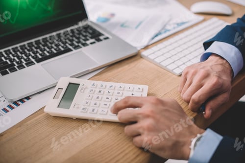 Preview: Cropped view of data analyst using calculator near laptop and papers on table