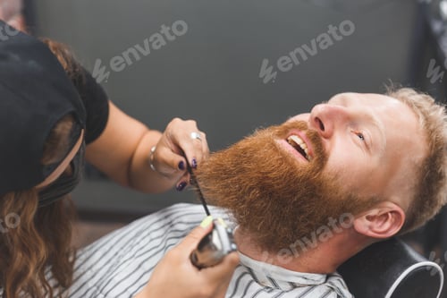 Preview: A man at a barbershop. Woman barber clipping beard and mustache. Barber woman in mask.