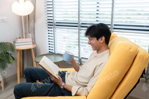 Preview: Young Man Relaxing in Modern Living Room, Reading a Book and Enjoying a Hot Beverage on a