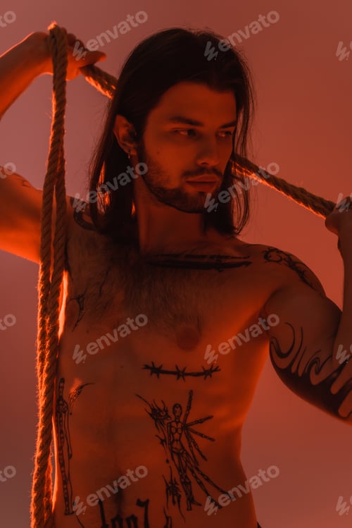 Preview: Young tattooed and shirtless model posing with rope isolated on red