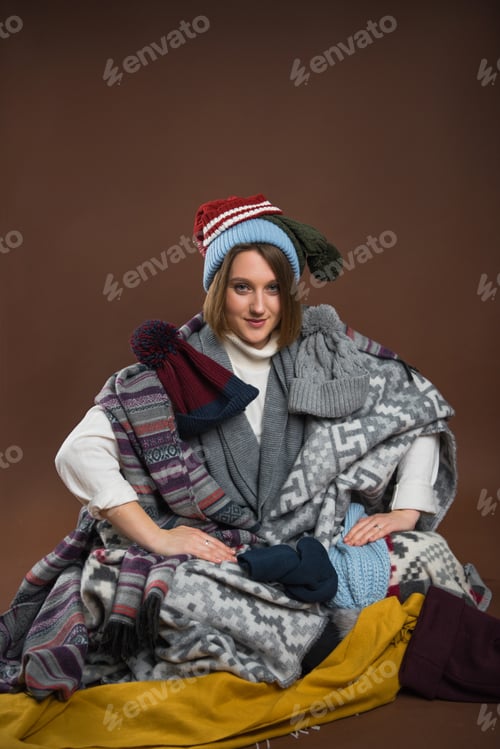 Preview: Woman sitting wrapped in blankets with winter hats and looking at camera isolated on brown