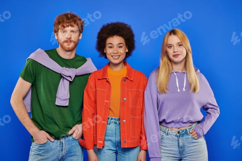 Preview: polyamory concept, cheerful multicultural women and redhead man looking at camera on blue