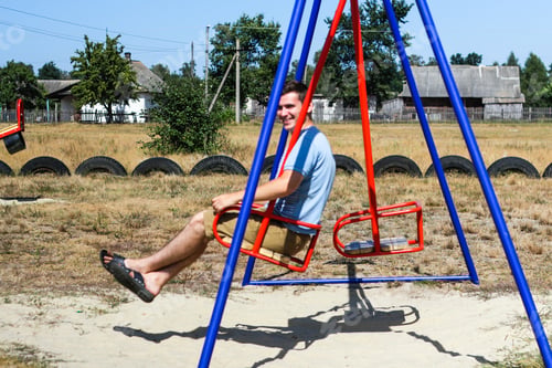 Preview: smiling young man swinging on swing on playground. Countryside area. Bright blue and red swing.