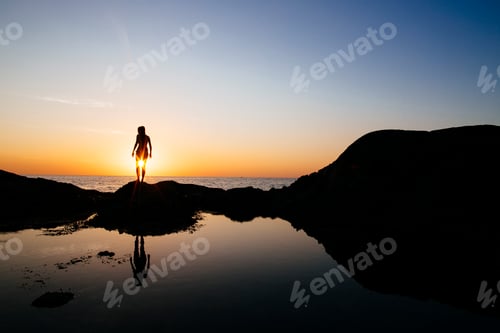 Preview: Silhouette of woman standing on a rock by the ocean at sunset.
