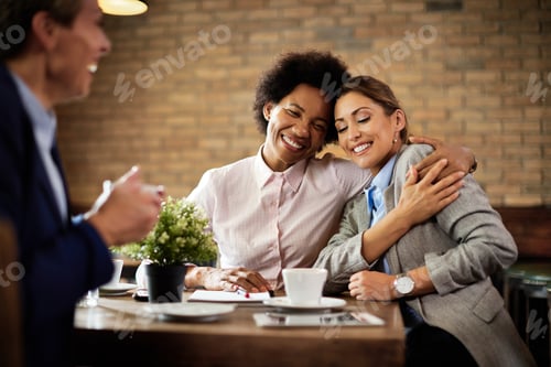 Preview: Happy female colleagues embracing with eyes closed while having coffee break in a cafe.