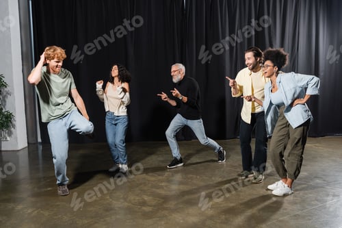 Preview: cheerful multicultural actors pointing at redhead man rehearsing on stage in theater
